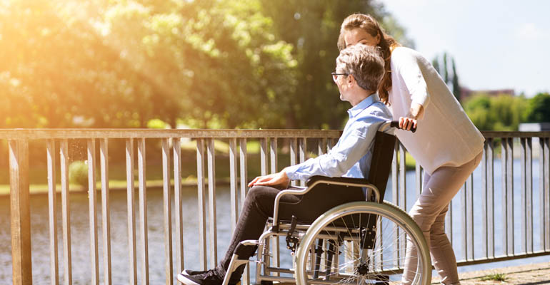 Foto på en man i rullstol och en kvinna som står på en bro och tittar ut över en älv.